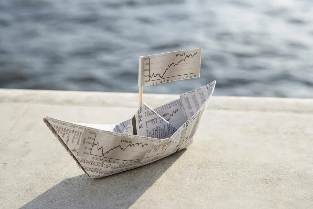 A newspaper paper boat with a stock-chart flag by the water